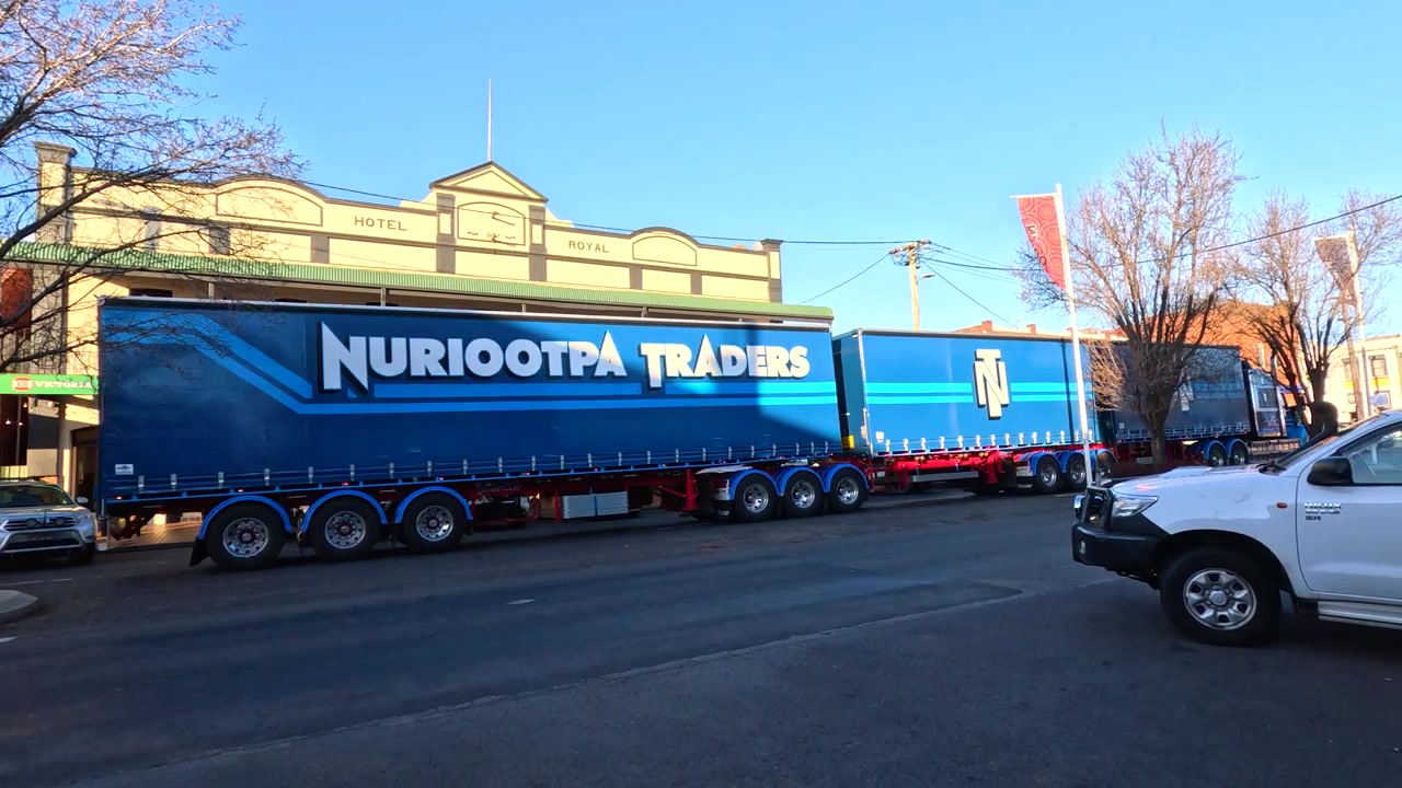 A blue road train drives through a quiet Australian town past historic buildings at sunset