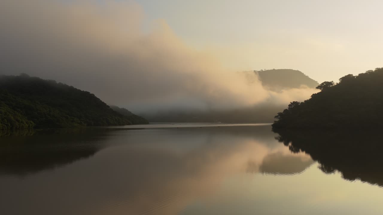 Breathtaking aerial view of misty Presa EL Carrizo in Jalisco at sunrise. Golden light reflects on calm waters, surrounded by lush greenery. Captivating, tranquil atmosphere