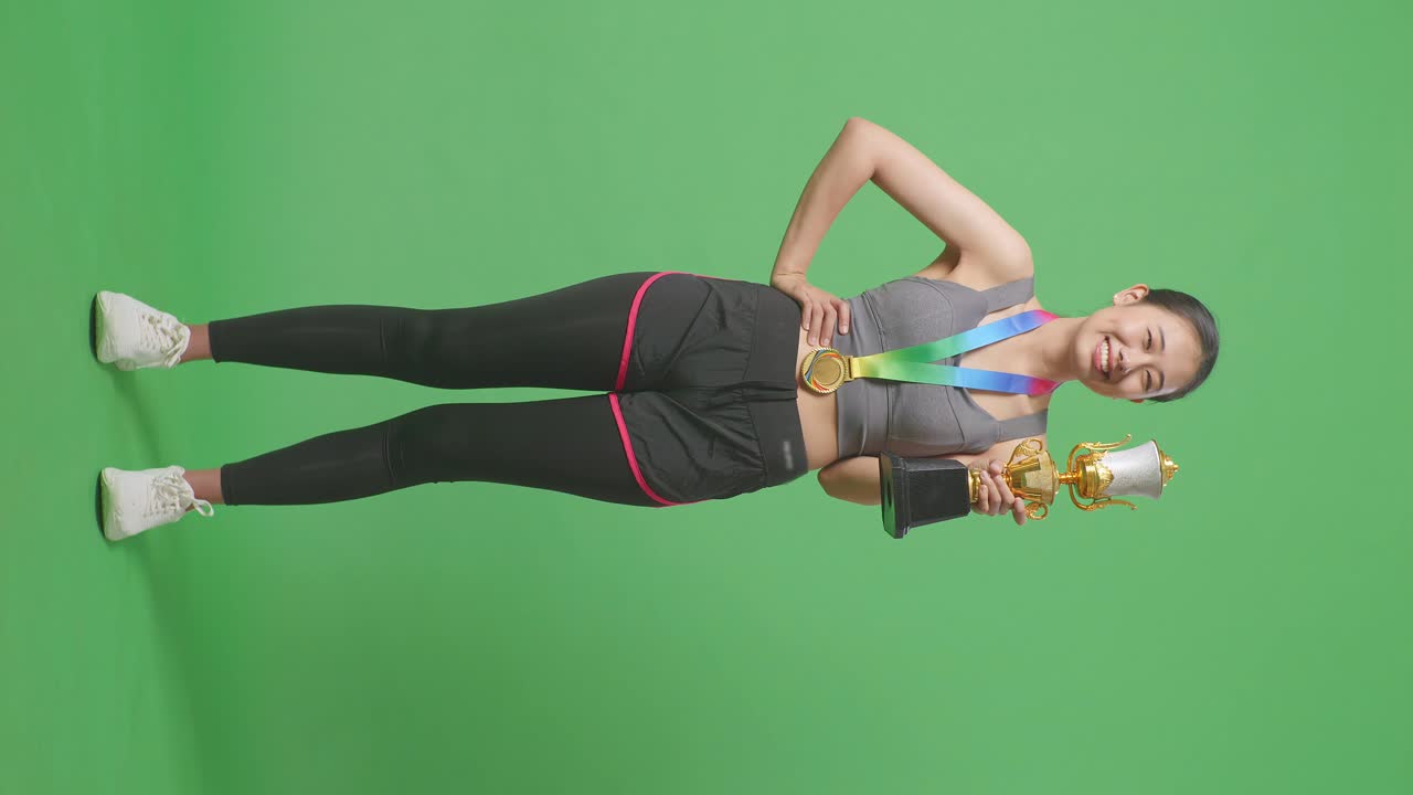 Donna asiatica a figura intera con una medaglia d'oro e un trofeo che sorride alla telecamera con un braccio sul fianco, in posa come prima classificata su sfondo green screen in studio.