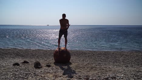 man doing yoga exercise outdoors near to an old rusty floating marine mine on the beach with rocky shore and sea background. healthy lifestyle, pollution, nature protection, war and peace concept