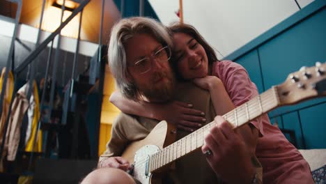 una rubia con cabello largo en gafas con barba toca la guitarra eléctrica, una chica morena abraza sus hombros y escucha al tipo tocando en una habitación acogedora