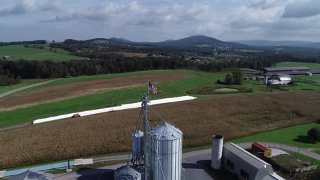 vista aérea alejándose de la bandera americana en la parte superior del silo de grano con campos de maíz y montañas en la distancia