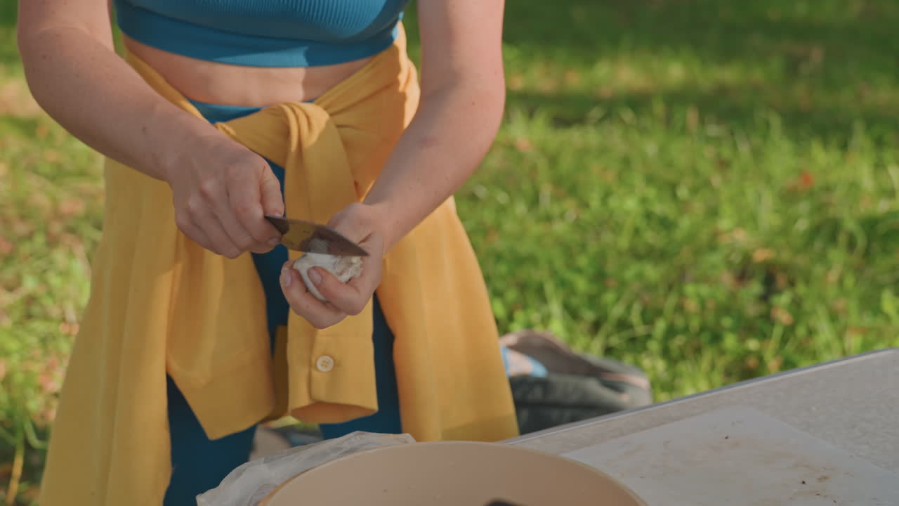Donna che pela aglio e cipolla su un tavolo da campeggio all'aperto, primo piano delle mani che usano coltello e pelapatate, luce solare che filtra tra gli alberi, ciotola da picnic con cipolla affettata, movimenti ritmici e attenti, campeggio estivo intimo.