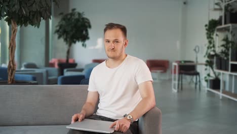 young man in the business center lobby sits down, closes his laptop, folds his hands, looks into the camera, and smiles