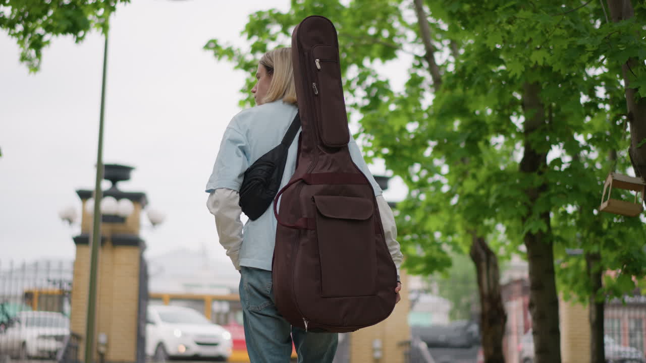 chitarrista bianco che si allontana sotto gli alberi sul sentiero di un parco urbano, con custodia imbottita per chitarra sulla schiena, giacca di jeans, vista posteriore spontanea, luce nuvolosa, atmosfera tranquilla e riflessiva, clip video di viaggio