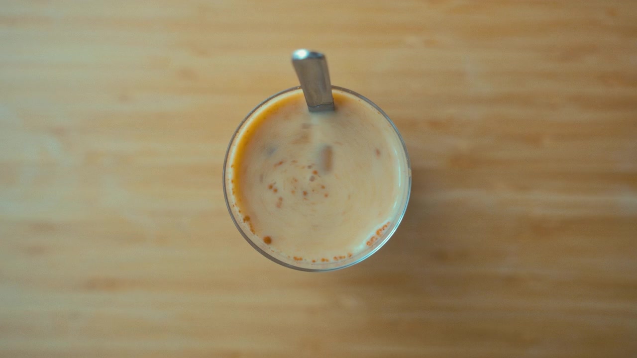Primo piano diurno di una mano che mescola un chai latte freddo con latte d'avena in un bicchiere con cannuccia su una superficie di legno.