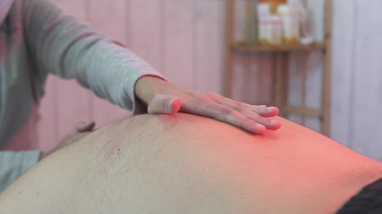Uomo adulto caucasico che riceve un trattamento di massaggio a infrarossi a luce rossa per il mal di schiena da una fisioterapista, senza primo piano del viso, al rallentatore con movimento.