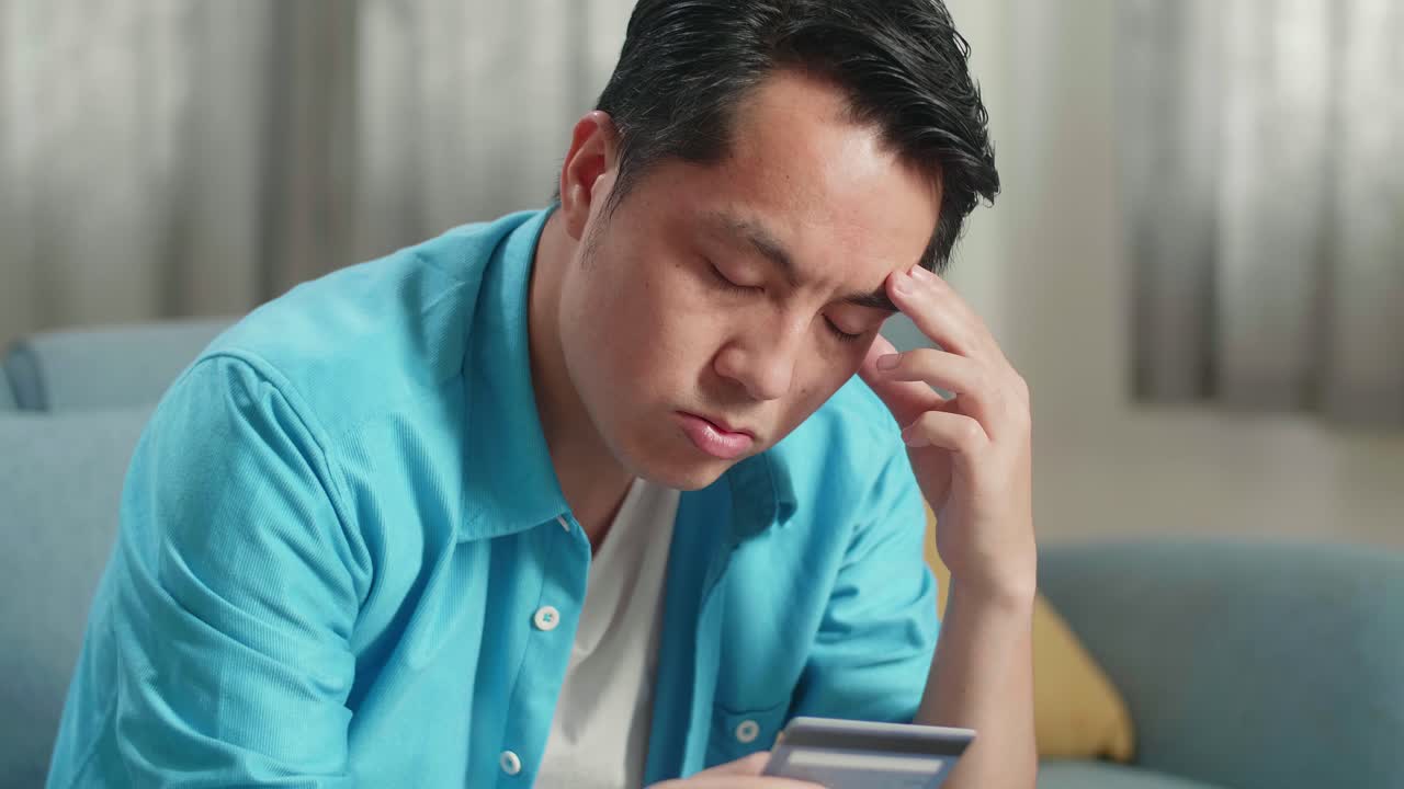 노트북을 들고 있는 아시아인 남자가 신용카드를 보고 재정 문제로 인해 두통을 고 있다.
