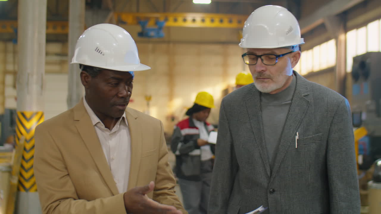 Multiethnic Businessmen in Hardhats Walking and Speaking in Factory