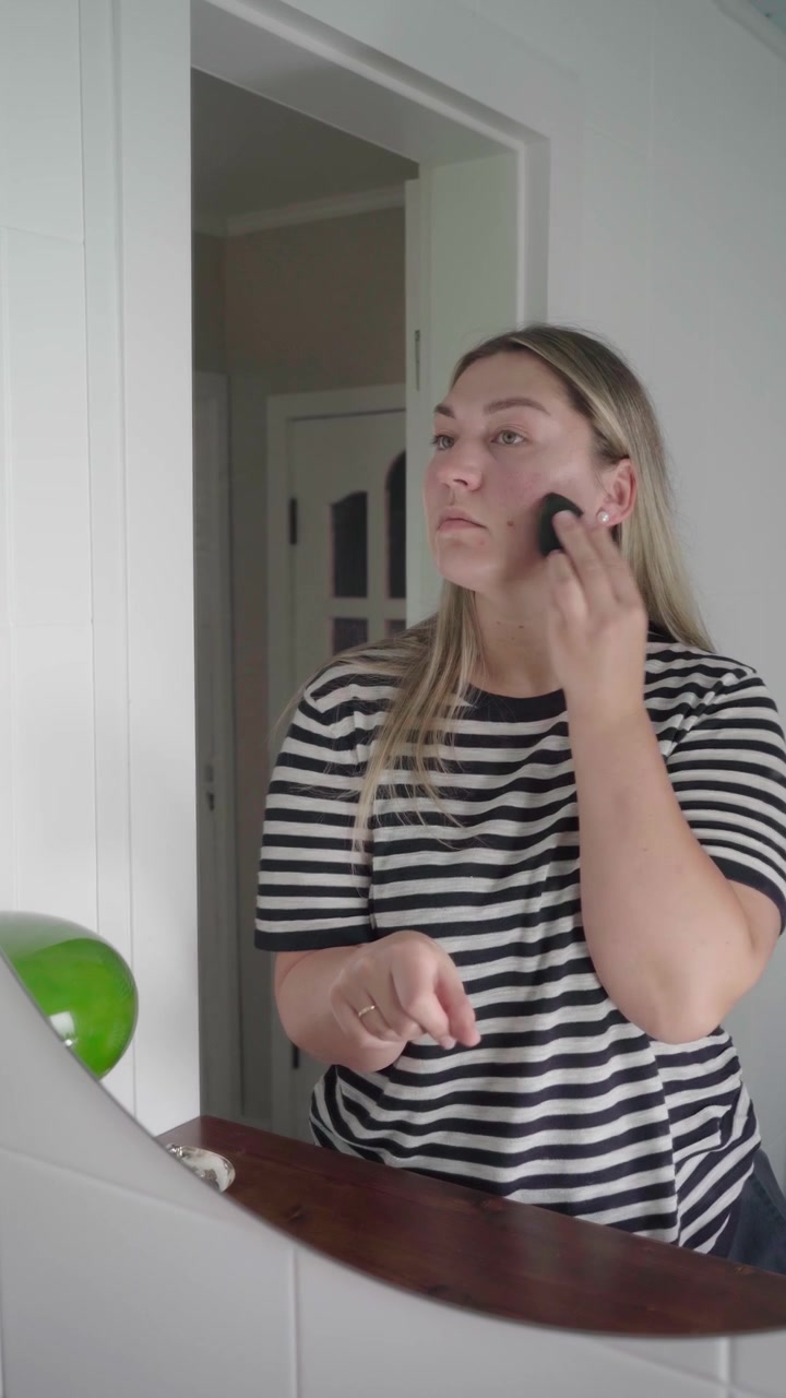 Woman in striped shirt dabs liquid foundation from her hand onto her face with a makeup sponge while facing the mirror.