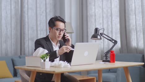 아시아 사업가 가 집 의 거실 에서 노트북 을 사용 하면서 온도계 를 들고 전화 를 하고 있다