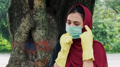 retrato de una hermosa joven afgana de cabello negro siguiendo la precaución y usando una máscara médica protectora y guantes y parada afuera cerca del árbol