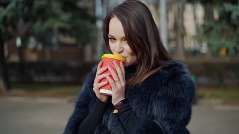 long-haired girl with plastic cup outdoors. attractive young woman in fur coat enjoys drinking coffee in the city park. slow motion.