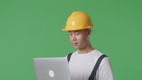 Close Up Of Side View Asian Man Painter Wearing Safety Helmet Using A Laptop While Standing In The Green Screen Background Studio