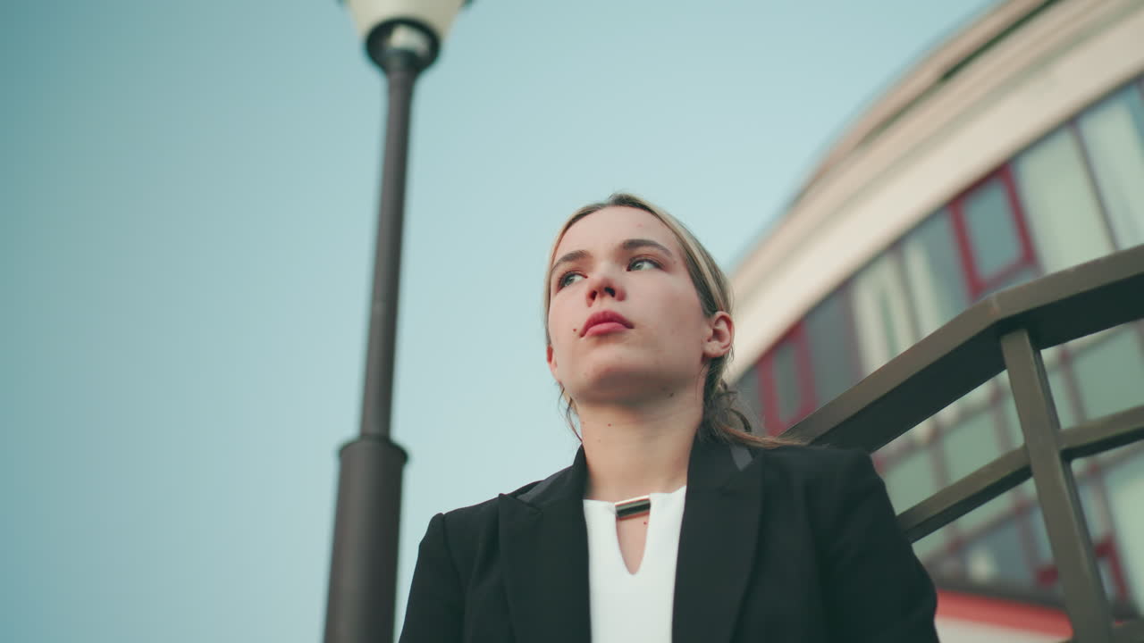 Worried woman in professional outfit looks tired and lost in thought with iron railing behind her and modern glass building in background