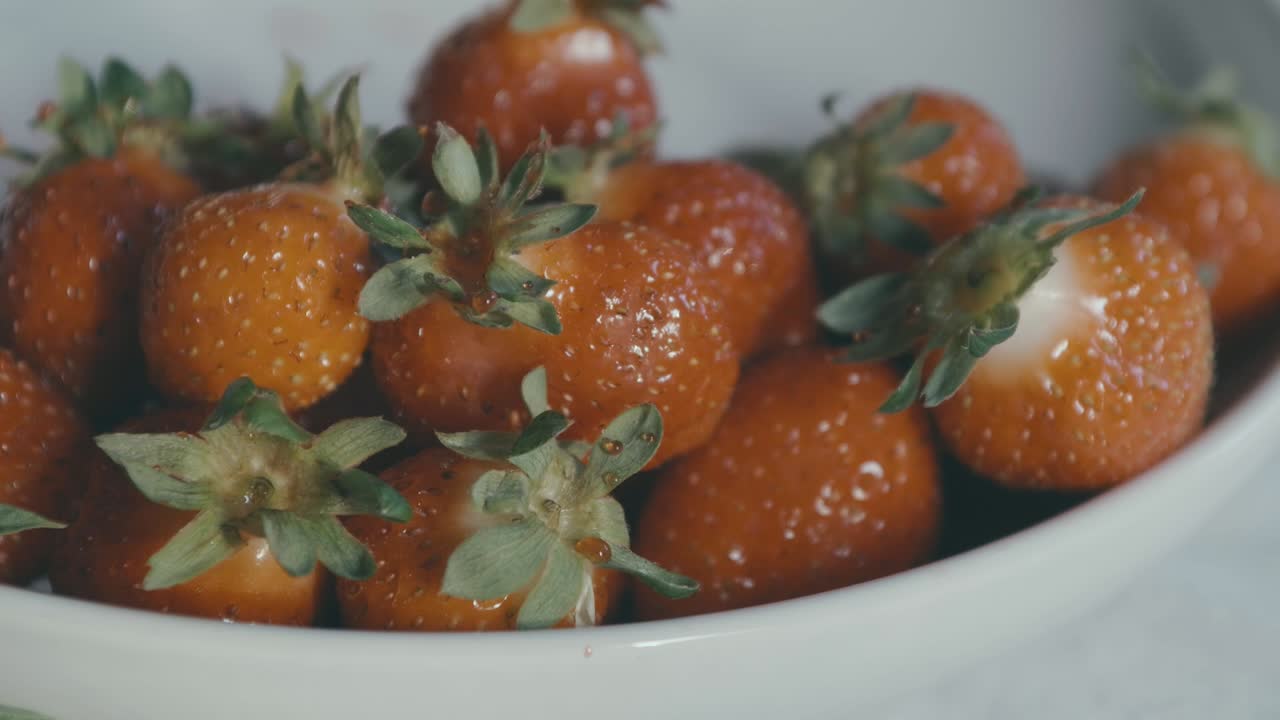 Una mano prende una fragola rossa da una ciotola piena di frutti rossi, la luce è naturale e il movimento è al rallentatore