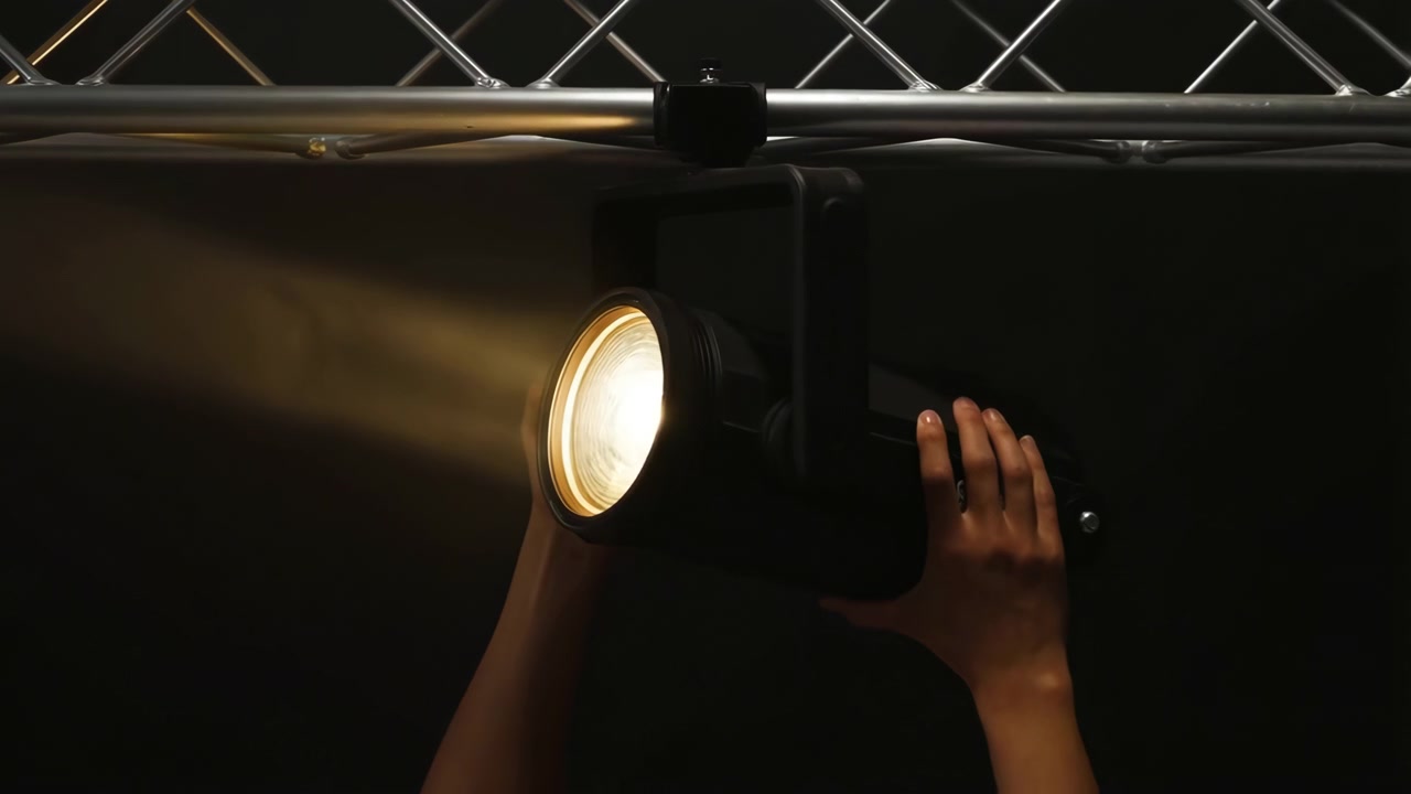 Vista cercana de dos manos ajustando un foco de escenario iluminado montado en una estructura metálica contra un telón de fondo negro intenso, mostrando el control manual de la iluminación, el mantenimiento y la preparación atmosférica de un concierto.