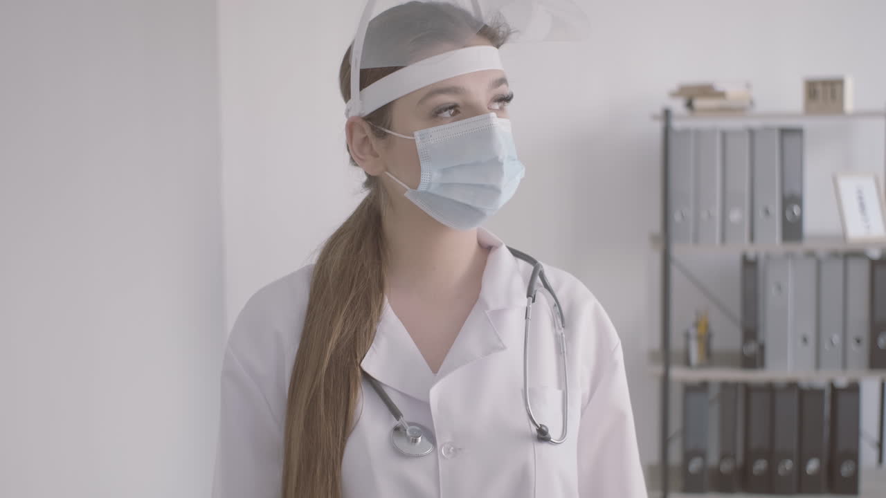 doctora pelirroja con bata blanca usando máscara médica y protección de pantalla facial mirando la cámara