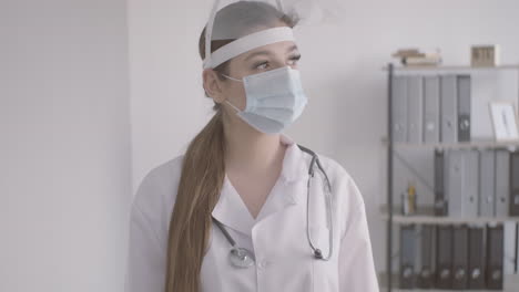 redhead doctor woman in white coat wearing medical mask and facial screen protection looking at the camera