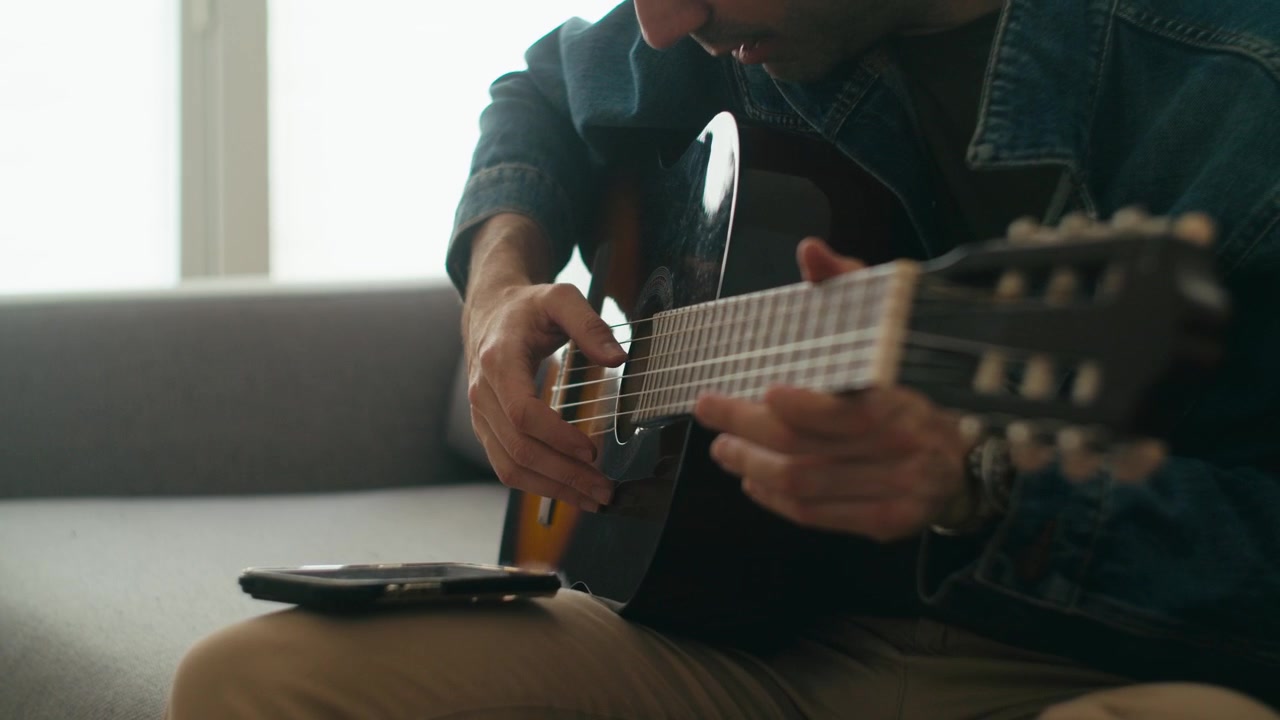 Un primo piano di un uomo con una giacca di jeans seduto su un divano, che suona una chitarra classica spagnola e con un'app per smartphone per lezioni di musica o accordatura
