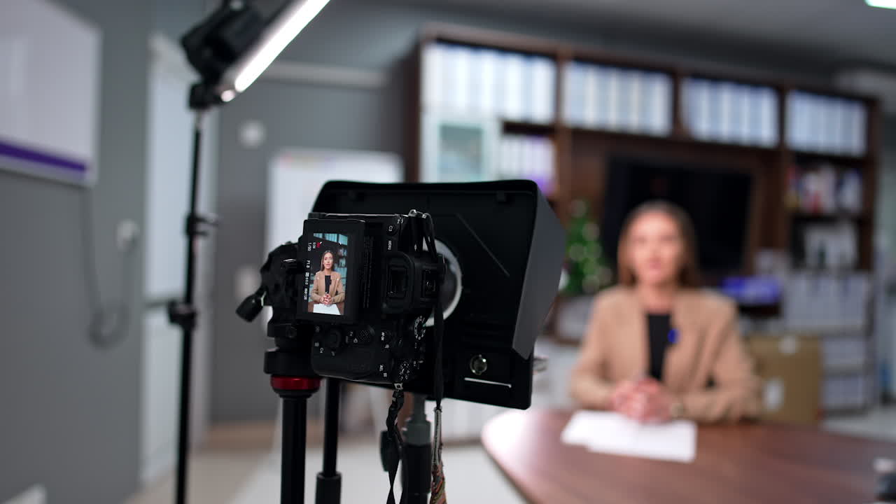 Blog content creation in the office. Woman wearing beige jacket records a video on professional camera. Blurred backdrop.