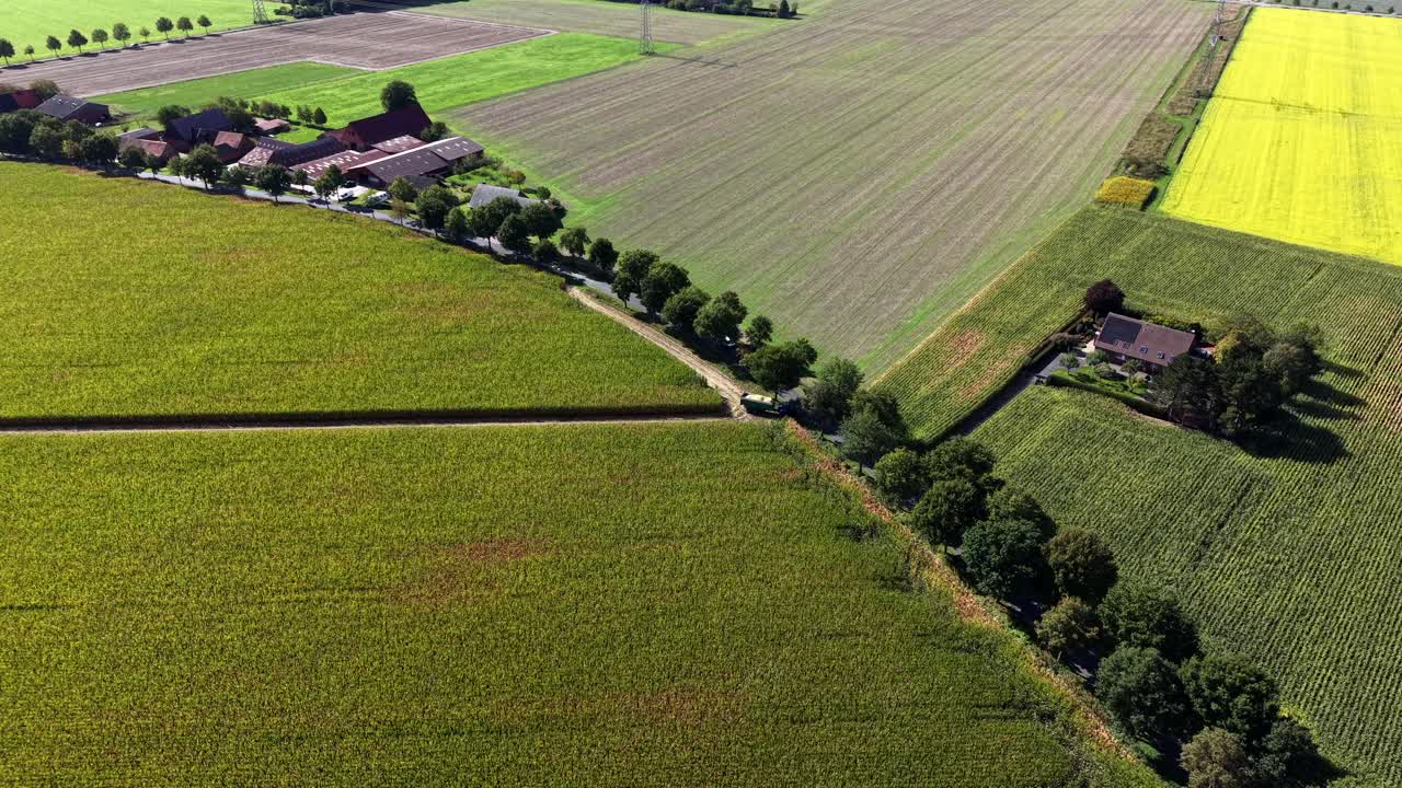 Tractor with trailer and harvested farm land fields of maize, canola and wheat in rural area of american town. Farmstead with houses, barn and garden- Aerial flyover shot. Sunny summer day
