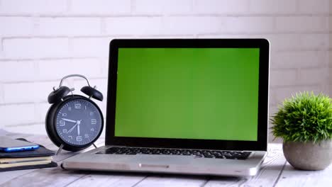 laptop with green screen and alarm clock on a wooden desk