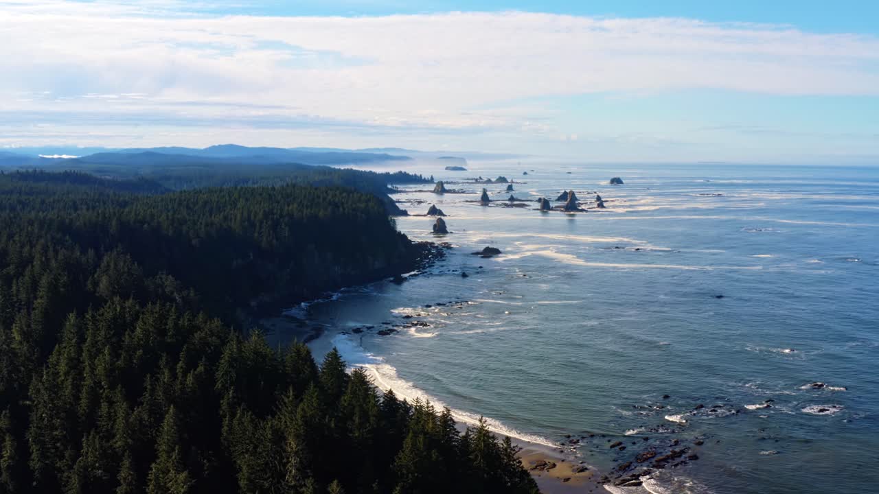 Stunning aerial drone time-lapse of the gorgeous Third Beach in Forks, Washington with large rock formations, surrounded by a pine tree forest on cliffs, and golden sand on a warm summer morning.