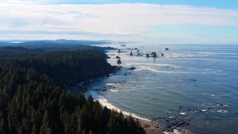 stunning aerial drone time-lapse of the gorgeous third beach in forks, washington with large rock formations, surrounded by a pine tree forest on cliffs, and golden sand on a warm summer morning.