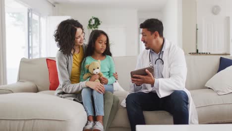 Hispanic-male-doctor-talking-to-mother-and-daughter-at-home,-all-wearing-face-masks