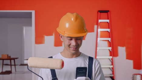 close up de un pintor asiático que usa un casco de seguridad sonriendo y usando un teléfono inteligente mientras está de pie pintando la pared en la habitación