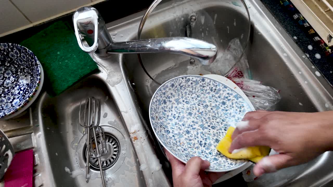 Housekeeper scrubbing dirty dishes in the kitchen sink with yellow sponge