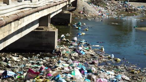 local river filled with plastics and other garbage near concrete bridge, static view