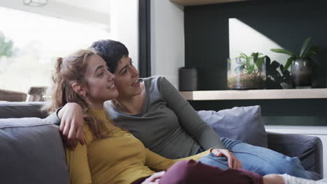 happy caucasian lesbian couple sitting on sofa, embracing and talking in sunny living room