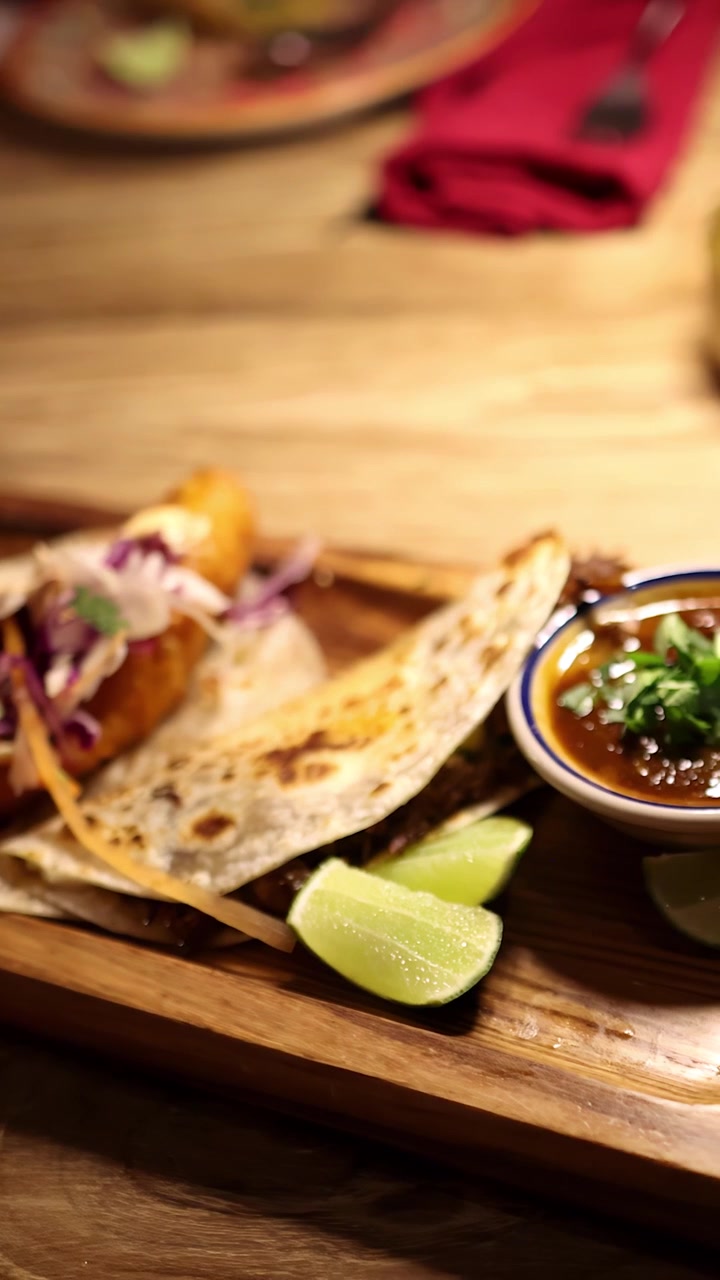 En person anretter forskellige grillede soft shell tacos på et træbræt under varm, stemningsfuld belysning, med synlige garniturer og dip i hyggelige spiseomgivelser.