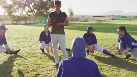 Diverse-group-of-female-baseball-players-with-coach,-warming-up-on-pitch,-squatting,-stretching-legs