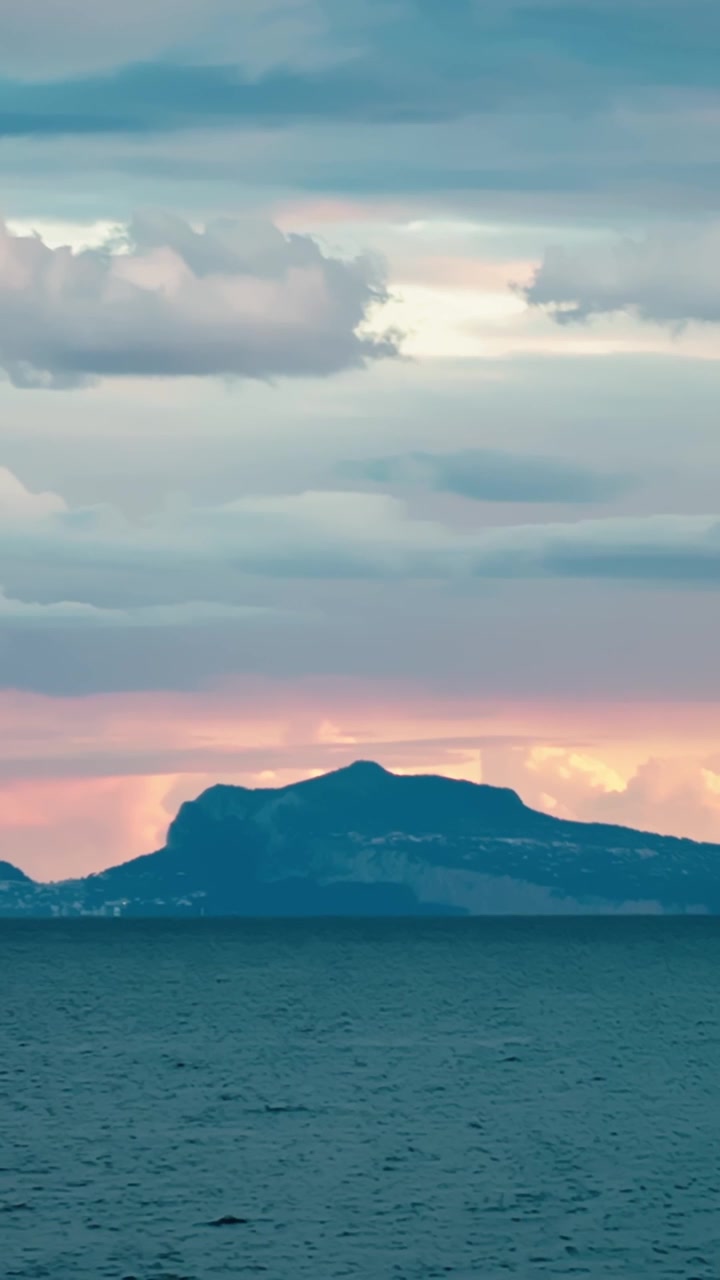 Silhouette dell'isola di Capri vista attraverso il Golfo di Napoli al tramonto, con acqua blu scuro, nuvole temporalesche stratificate e un orizzonte rosa tenue sul Mar Tirreno.