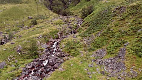 panning aerial shot, slowly panning across a close up of a fast flowing, steep rocky stream on the lush, green valley hillside of glen coe, scotland, united kingdom, europe