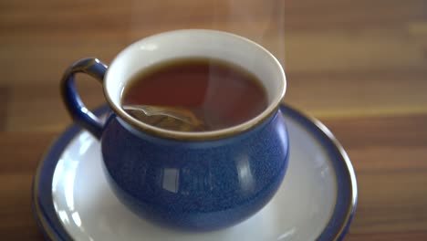 small bag of black tea bobbling in a steaming hot water in a blue tea cup on a wooden table into the kitchen