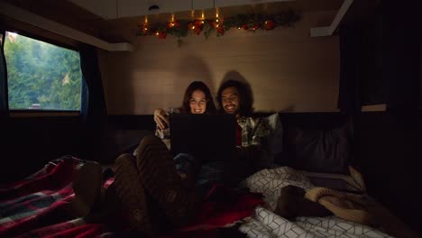 shooting in an atmospheric trailer with dim lighting, a happy brunette couple in plaid shirts lie in their shoes on the trailer sofa and watch a movie through a laptop near their camp outside the city in the summer