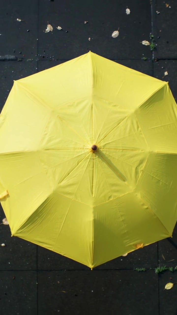 Video verticale: Ombrello giallo che ruota con pomello in legno su piastrelle scure mosso dalla brezza, foglie cadute.