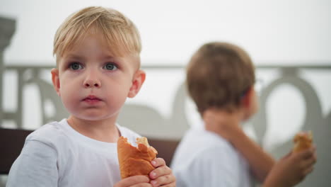 two siblings are seated while eating their snacks,the older brother looks around observantly, while the younger child appears contemplative, holding a sausage roll