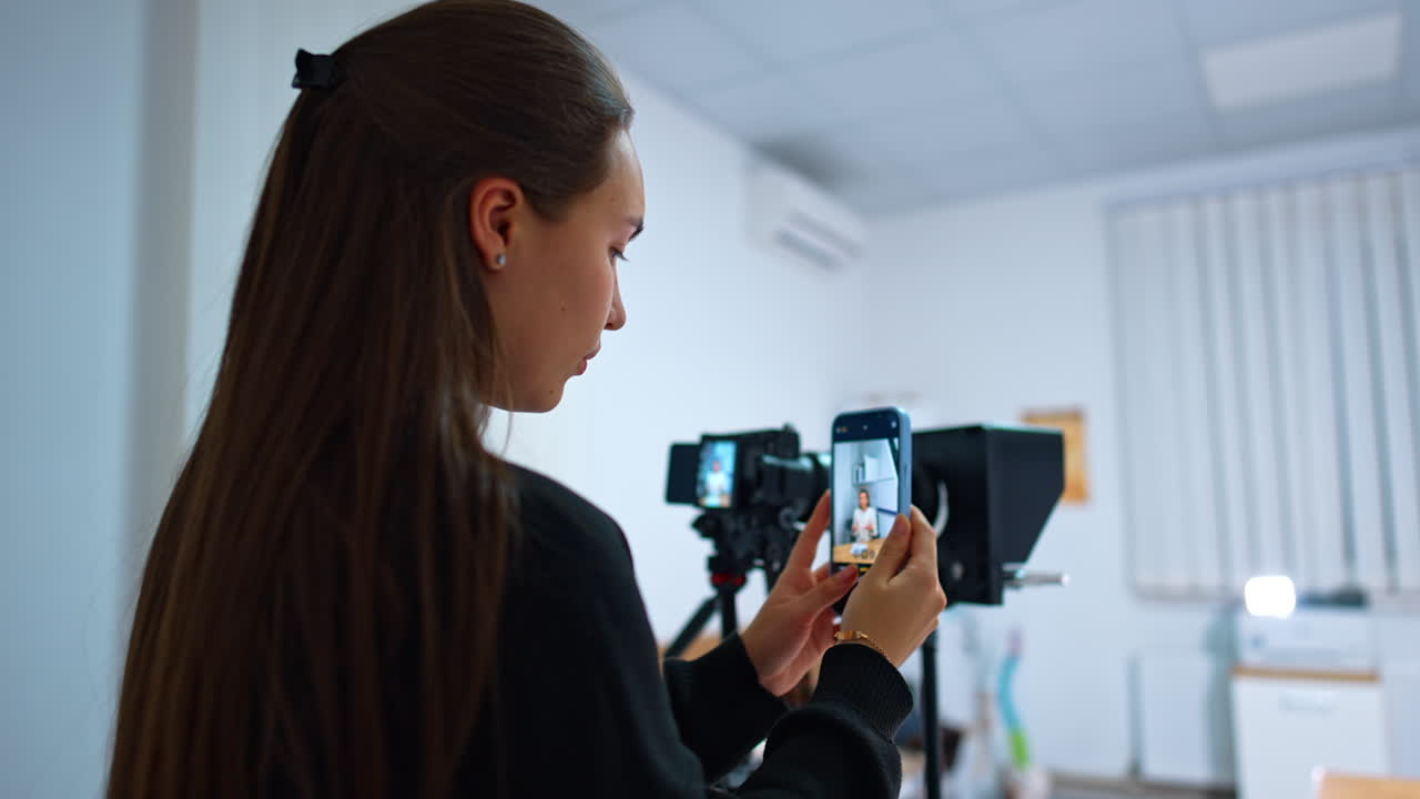 Long-haired brunette stands near the camera on tripod holding her phone. Girl takes video of a female speaker on her smartphone. Blurred backdrop.