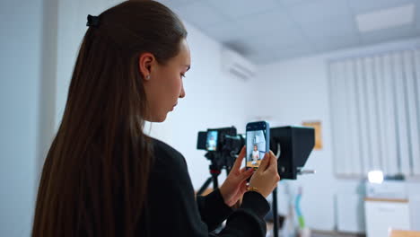 long-haired brunette stands near the camera on tripod holding her phone. girl takes video of a female speaker on her smartphone. blurred backdrop.