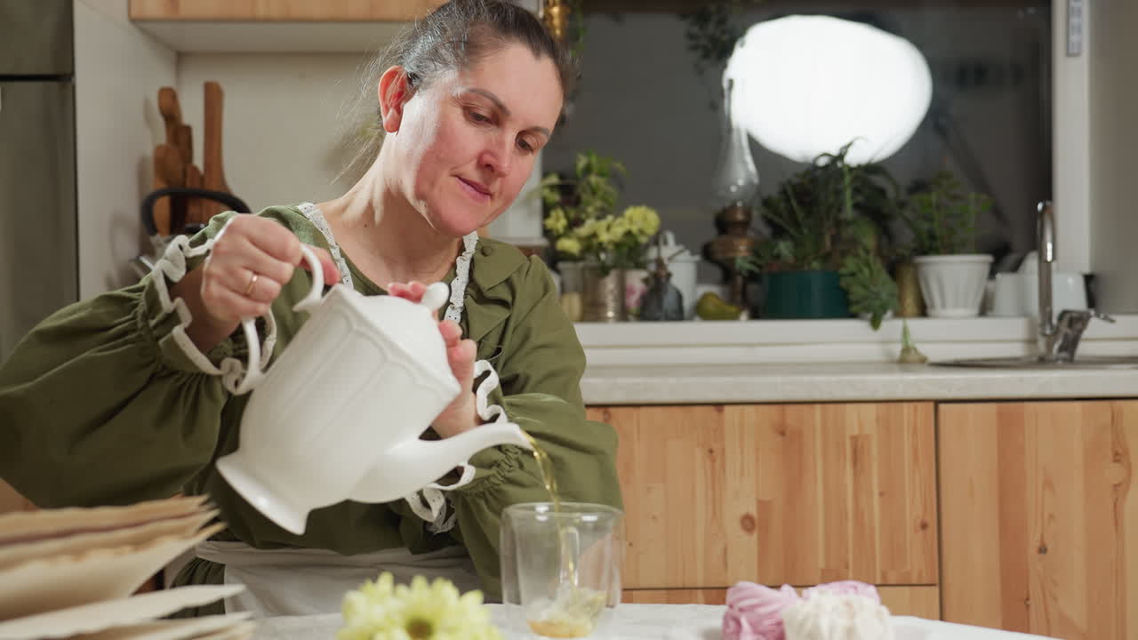 Mature woman wearing green dress and apron carefully pouring herbal tea from white ceramic teapot into clear glass cup while sitting at cozy kitchen table with flowers, pastries, and open recipe book