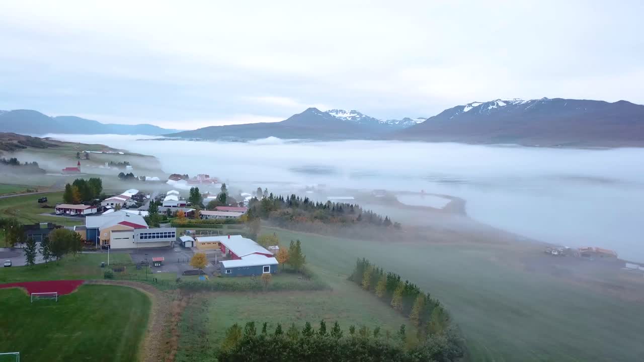 una toma panorámica de camiones de un fiordo muy temprano en la mañana con edificios y mucha niebla con montañas nevadas en el fondo y árboles verdes brillantes en primer plano