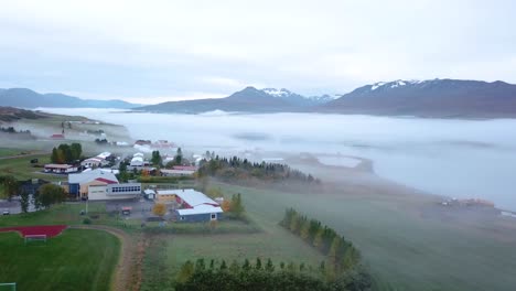 Una-Toma-Panorámica-De-Camiones-De-Un-Fiordo-Muy-Temprano-En-La-Mañana-Con-Edificios-Y-Mucha-Niebla-Con-Montañas-Nevadas-En-El-Fondo-Y-árboles-Verdes-Brillantes-En-Primer-Plano