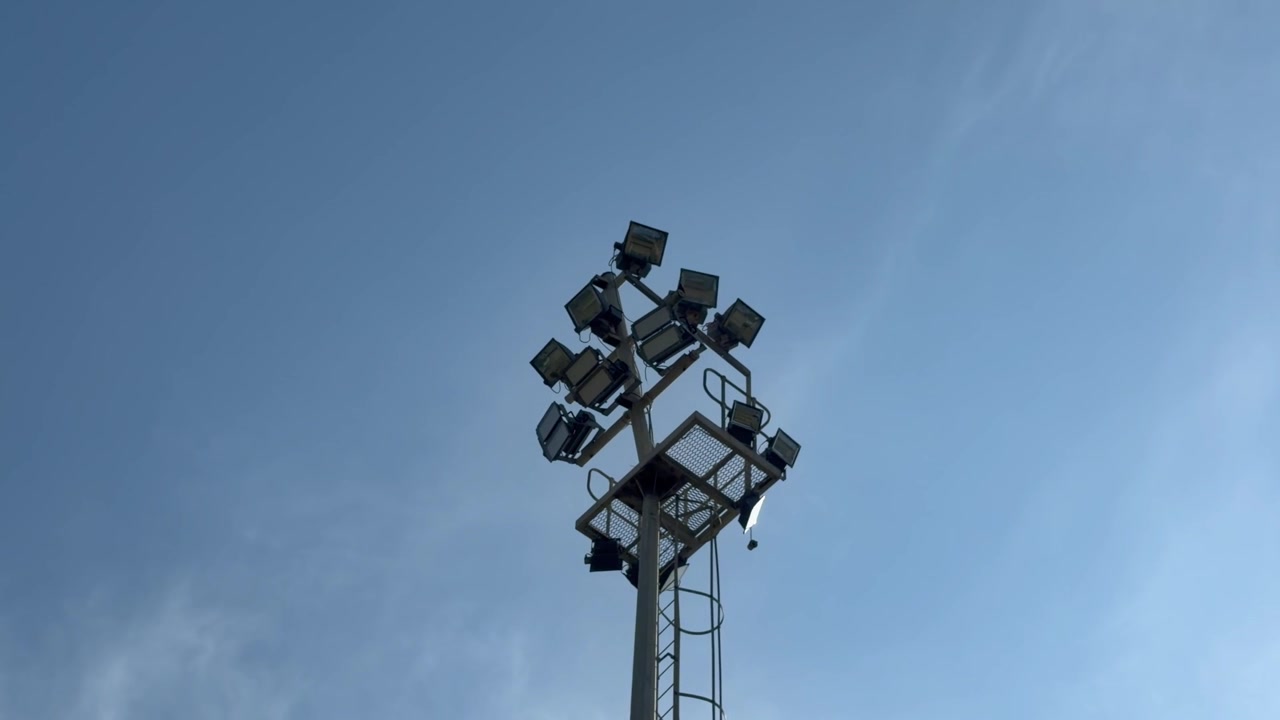 柔らかな青空を背景にそびえ立つスタジアムの照明ポール。その多数の大型投光器は、安全手すりと昇降はしごを備えた金属製整備台に取り付けられている