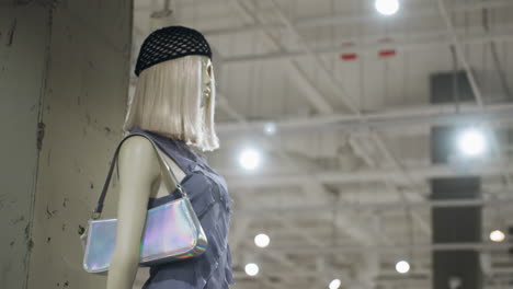 un maniquí en una tienda de ropa, vestido con una camiseta sin mangas y accesoriado con un bolso holográfico de moda. el maniquí tiene una peluca rubia y lleva un sombrero de red negra, luces brillantes de la tienda