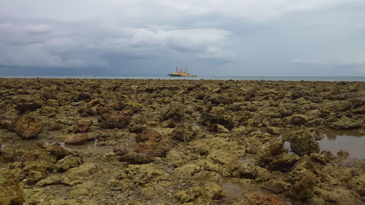 A fixed shot of Jagged rocks stretch toward the horizon where a smoke stacked ship drifts beneath heavy clouds of Mauban Port, Quezon Province Philippines
